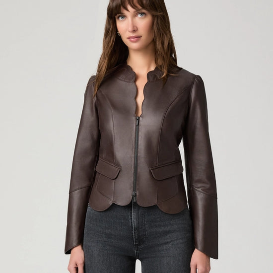 A woman modeling a dark brown leather jacket with a split collar and front zipper, paired with dark gray jeans, against a light gray studio background.