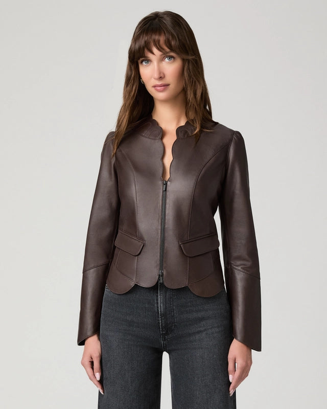 A woman modeling a dark brown leather jacket with a split collar and front zipper, paired with dark gray jeans, against a light gray studio background.