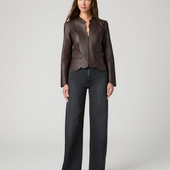 A woman modeling a dark brown leather jacket with a split neckline and front zipper, paired with dark gray wide-leg jeans, against a light gray studio background.