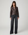 A woman modeling a dark brown leather jacket with a split neckline and front zipper, paired with dark gray wide-leg jeans, against a light gray studio background.