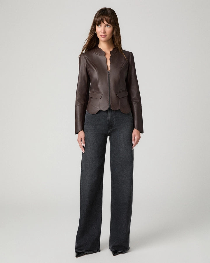 A woman modeling a dark brown leather jacket with a split neckline and front zipper, paired with dark gray wide-leg jeans, against a light gray studio background.