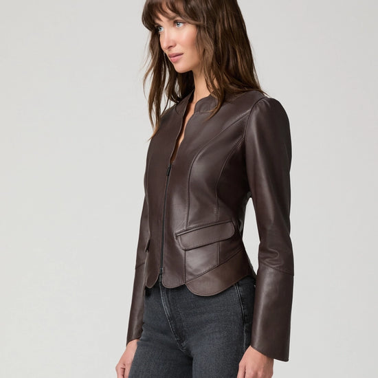 A woman wearing a dark brown leather jacket with structured shoulders and a front zipper, paired with dark gray jeans, photographed against a light gray studio background.