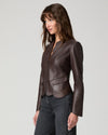 A woman wearing a dark brown leather jacket with structured shoulders and a front zipper, paired with dark gray jeans, photographed against a light gray studio background.