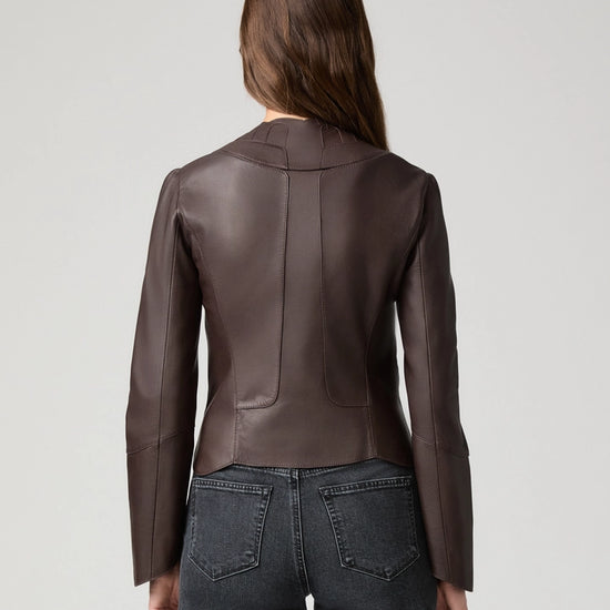 A woman viewed from the back wearing a fitted dark brown leather jacket and dark gray jeans against a light gray studio background.