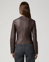 A woman viewed from the back wearing a fitted dark brown leather jacket and dark gray jeans against a light gray studio background.
