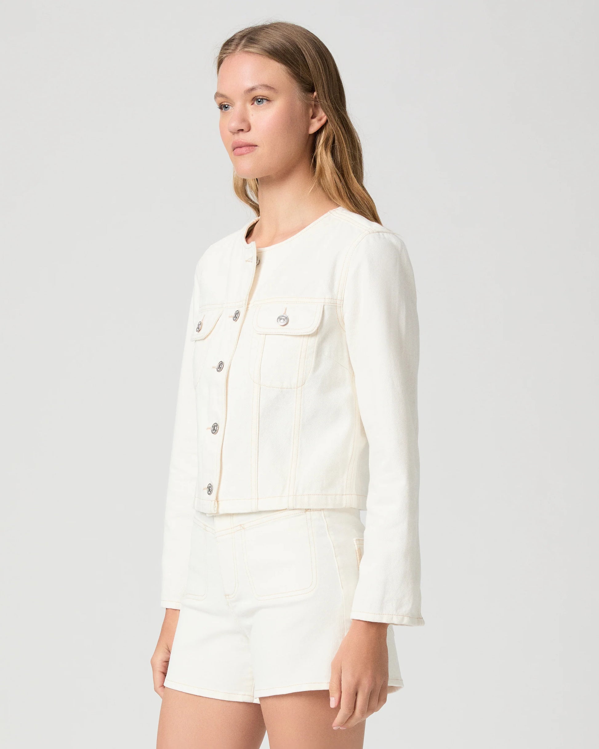 Woman wearing a cream Paige denim jacket and shorts on a plain background side view