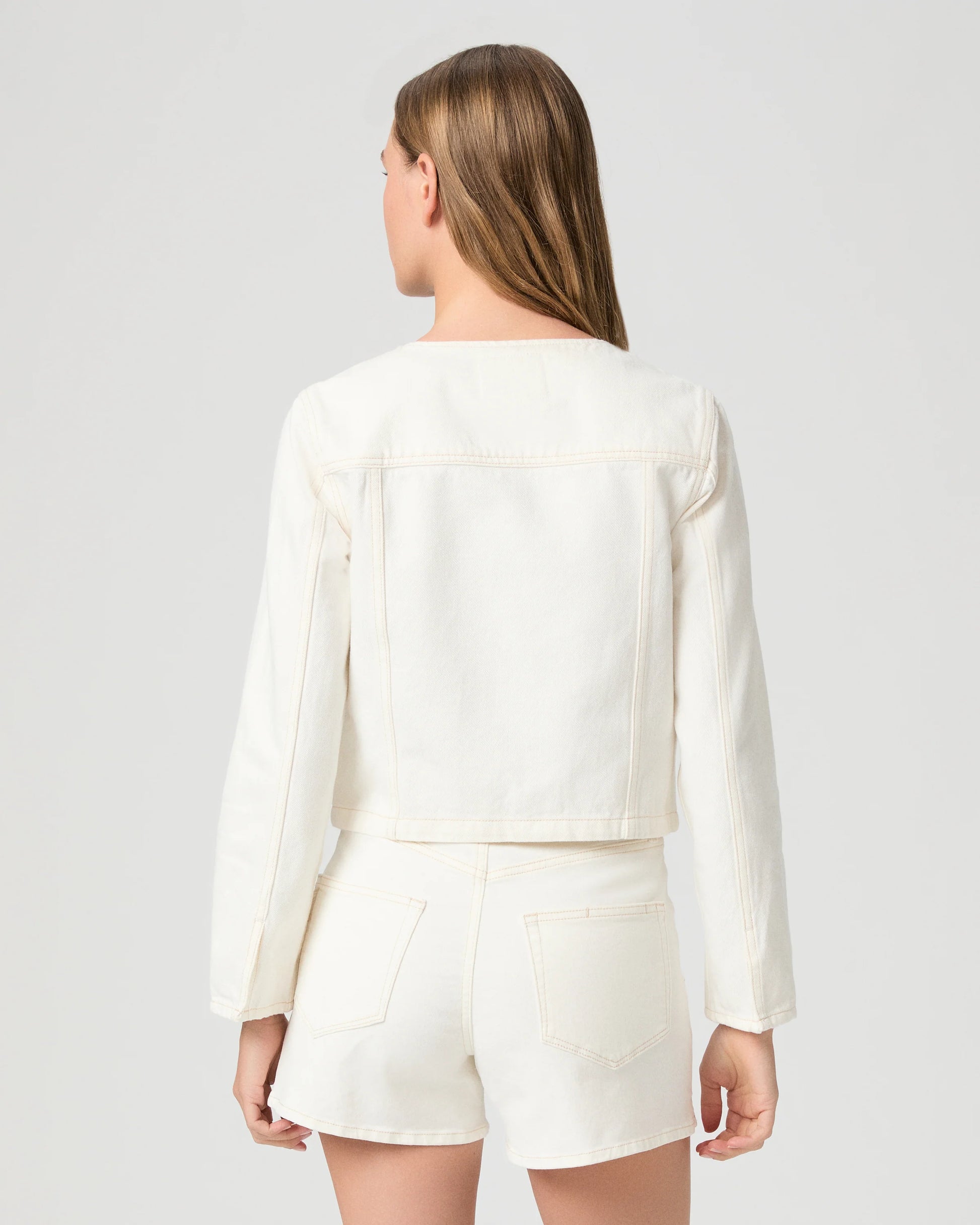 Woman wearing a cream Paige denim jacket and shorts on a plain background rear view
