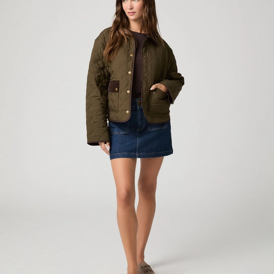 A woman modeling a cropped quilted jacket in olive green with brown contrast panels and gold-tone snap buttons, worn over a dark top and blue denim skirt, against a light gray studio background.