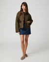 A woman modeling a cropped quilted jacket in olive green with brown contrast panels and gold-tone snap buttons, worn over a dark top and blue denim skirt, against a light gray studio background.