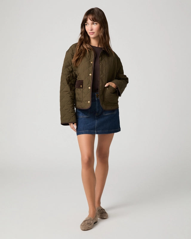 A woman modeling a cropped quilted jacket in olive green with brown contrast panels and gold-tone snap buttons, worn over a dark top and blue denim skirt, against a light gray studio background.