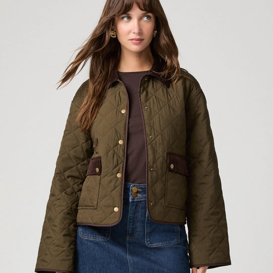 A woman modeling a quilted olive green jacket with brown trim, worn over a dark brown top and a blue denim mini skirt, against a light gray studio background.