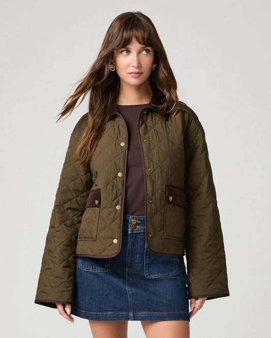 A woman modeling a quilted olive green jacket with brown trim, worn over a dark brown top and a blue denim mini skirt, against a light gray studio background.