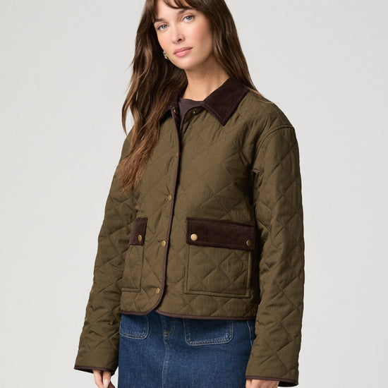 A woman modeling a quilted olive green jacket with dark brown collar and pocket flap details, paired with a blue denim skirt, against a light gray studio background.