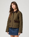 A woman modeling a quilted olive green jacket with dark brown collar and pocket flap details, paired with a blue denim skirt, against a light gray studio background.