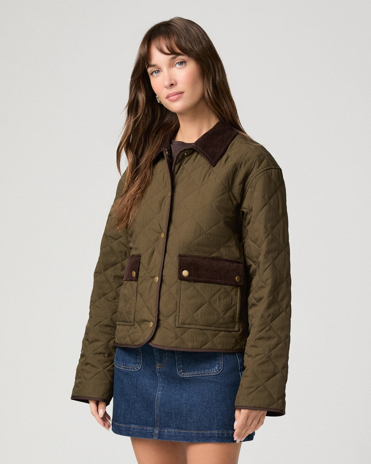 A woman modeling a quilted olive green jacket with dark brown collar and pocket flap details, paired with a blue denim skirt, against a light gray studio background.