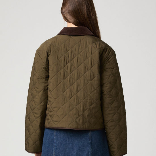 A woman shown from the back wearing a quilted olive green jacket with a dark brown collar and a blue denim skirt, against a light gray studio background.