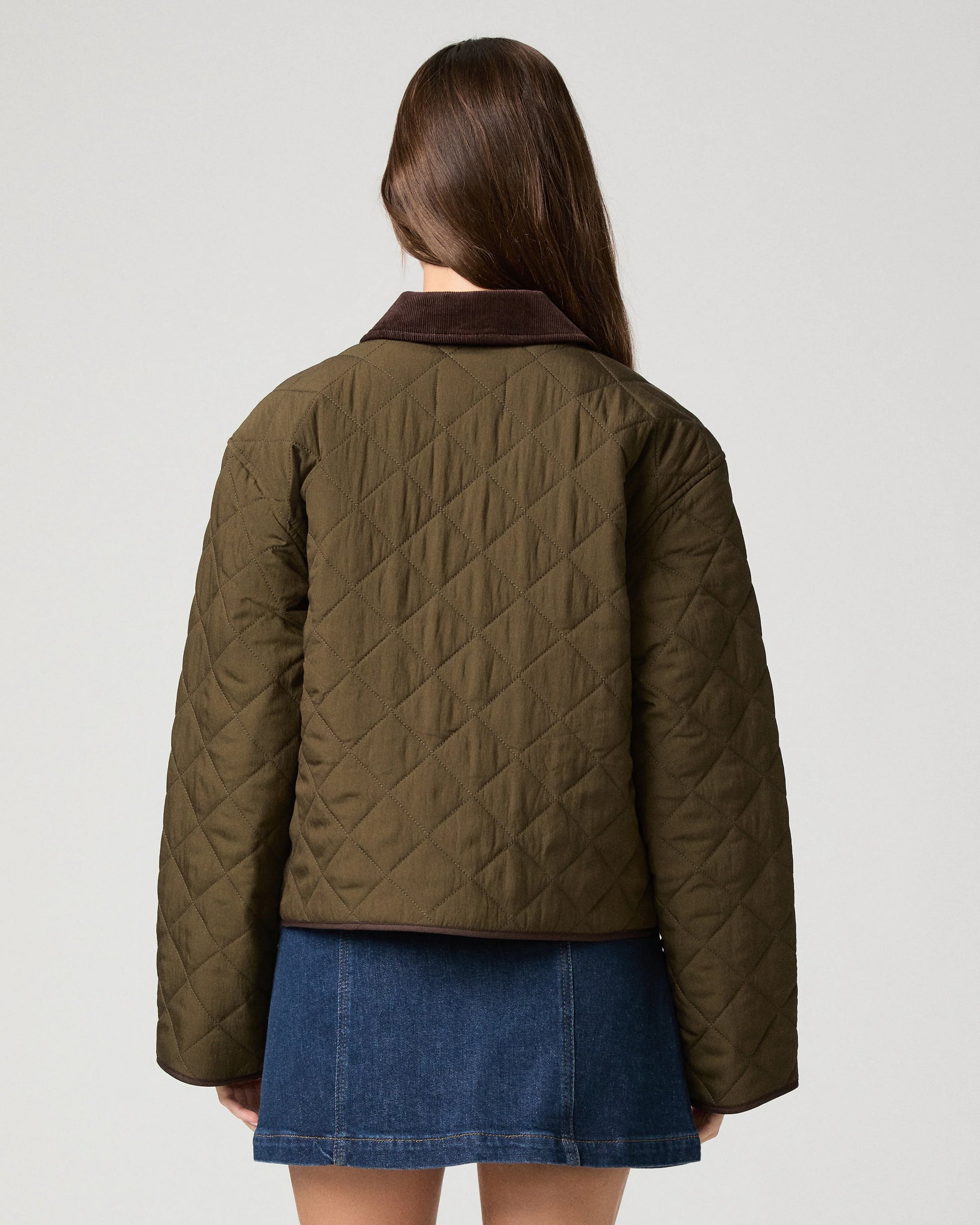 A woman shown from the back wearing a quilted olive green jacket with a dark brown collar and a blue denim skirt, against a light gray studio background.