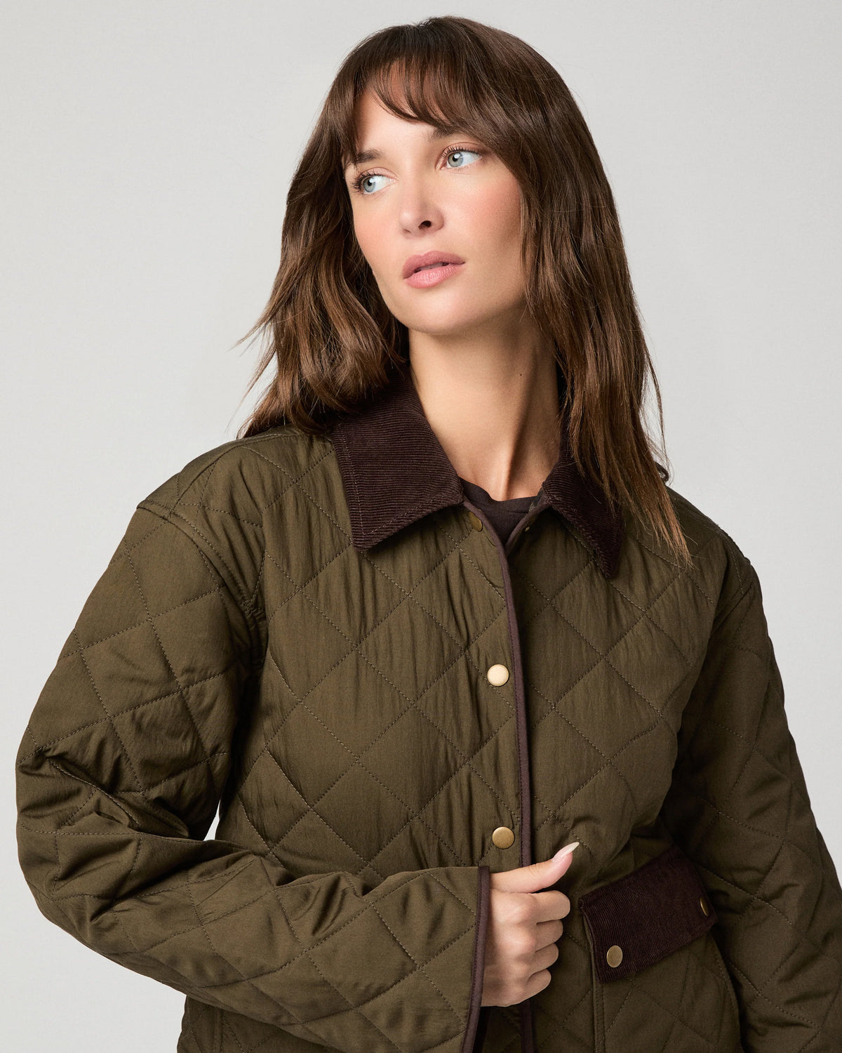 A woman wearing an olive green diamond-quilted jacket with a dark brown corduroy collar and brown button details, photographed against a neutral light gray background.