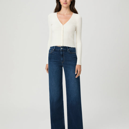A woman modeling a white long-sleeve V-neck knit top and dark blue wide-leg jeans against a light gray studio background.