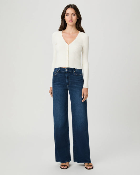 A woman modeling a white long-sleeve V-neck knit top and dark blue wide-leg jeans against a light gray studio background.