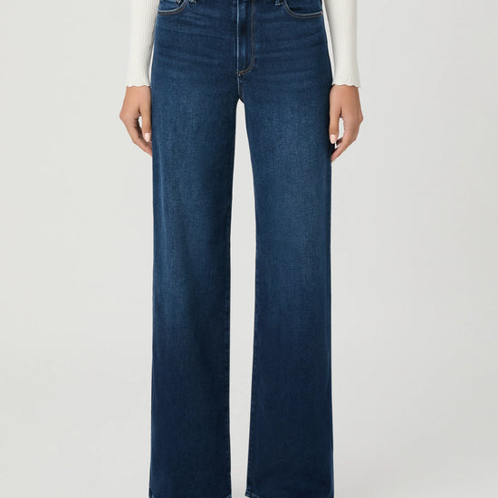 A model wearing dark blue wide-leg denim jeans, a white ribbed long-sleeve top, and brown sandals, shown from the waist down against a light gray studio background.