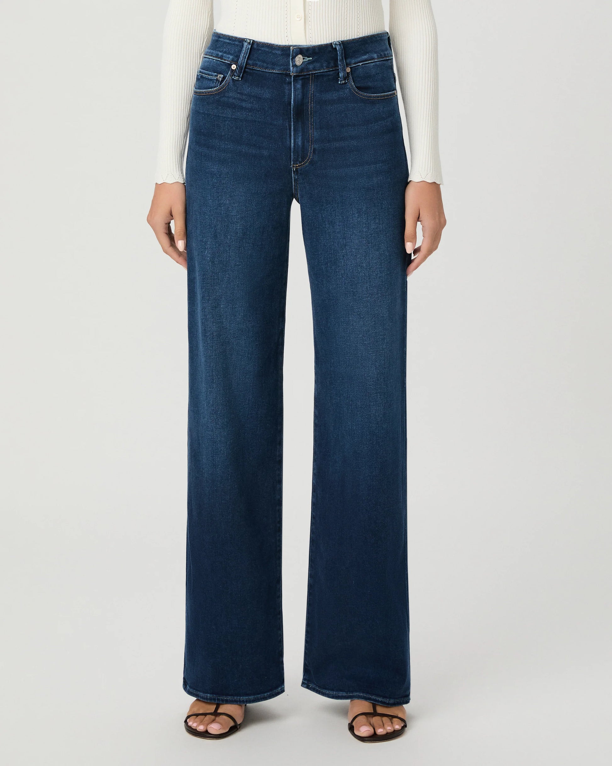 A model wearing dark blue wide-leg denim jeans, a white ribbed long-sleeve top, and brown sandals, shown from the waist down against a light gray studio background.