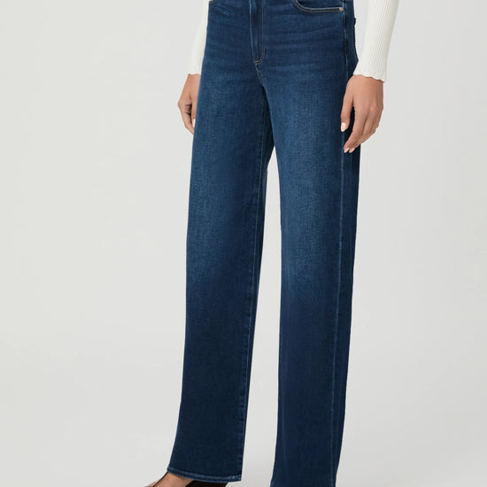 A model wearing dark blue wide-leg jeans, a white ribbed long-sleeve top, and brown flat sandals, shown from the waist down against a light gray studio background.