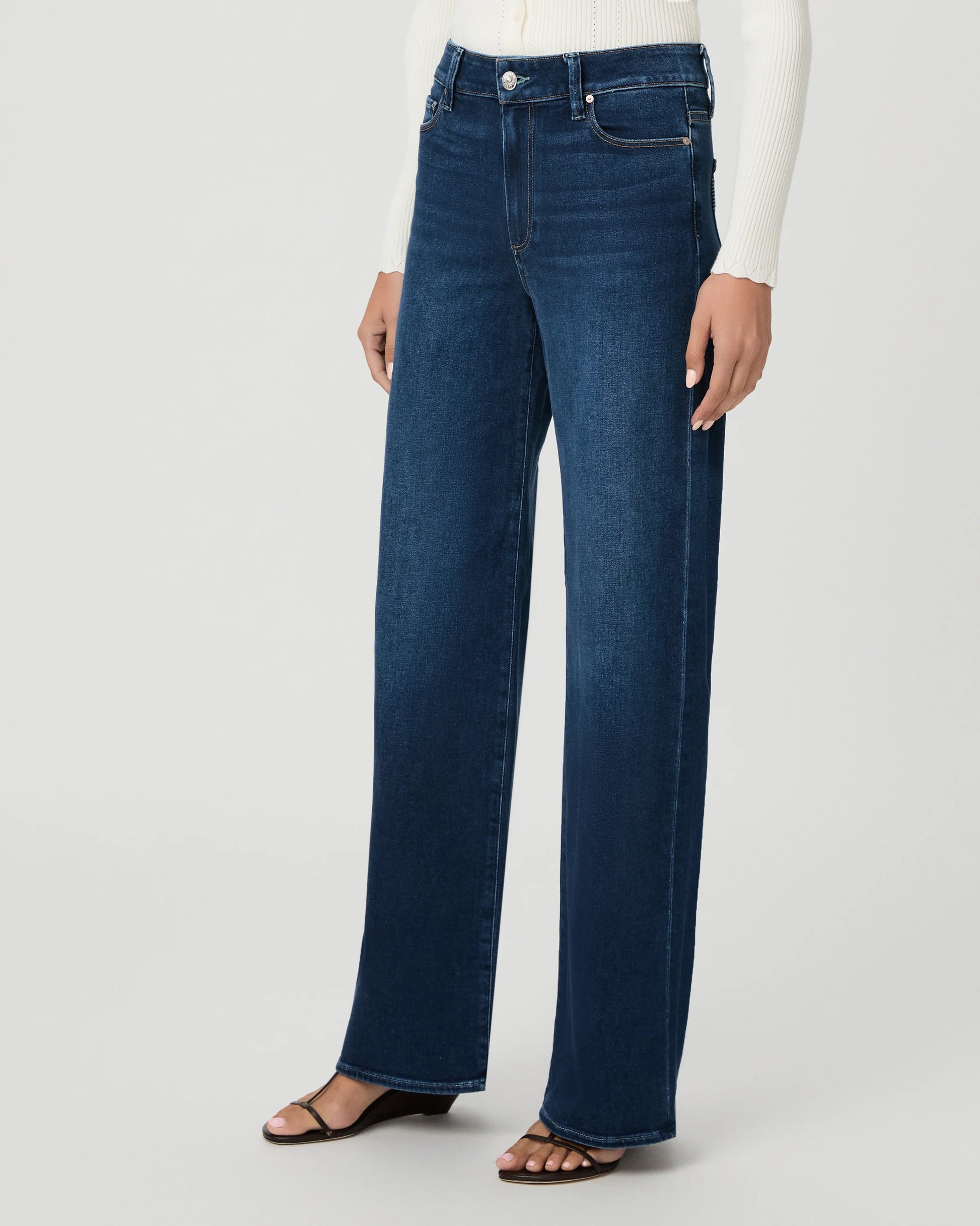 A model wearing dark blue wide-leg jeans, a white ribbed long-sleeve top, and brown flat sandals, shown from the waist down against a light gray studio background.
