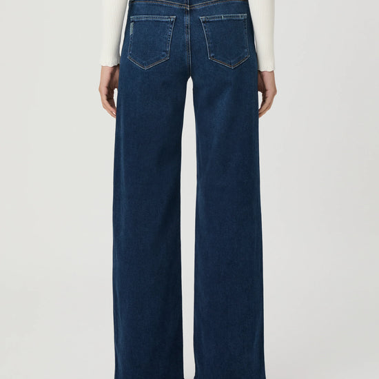 Back view of a model wearing dark blue wide-leg denim jeans with a white long-sleeve top, shown against a light gray studio background.