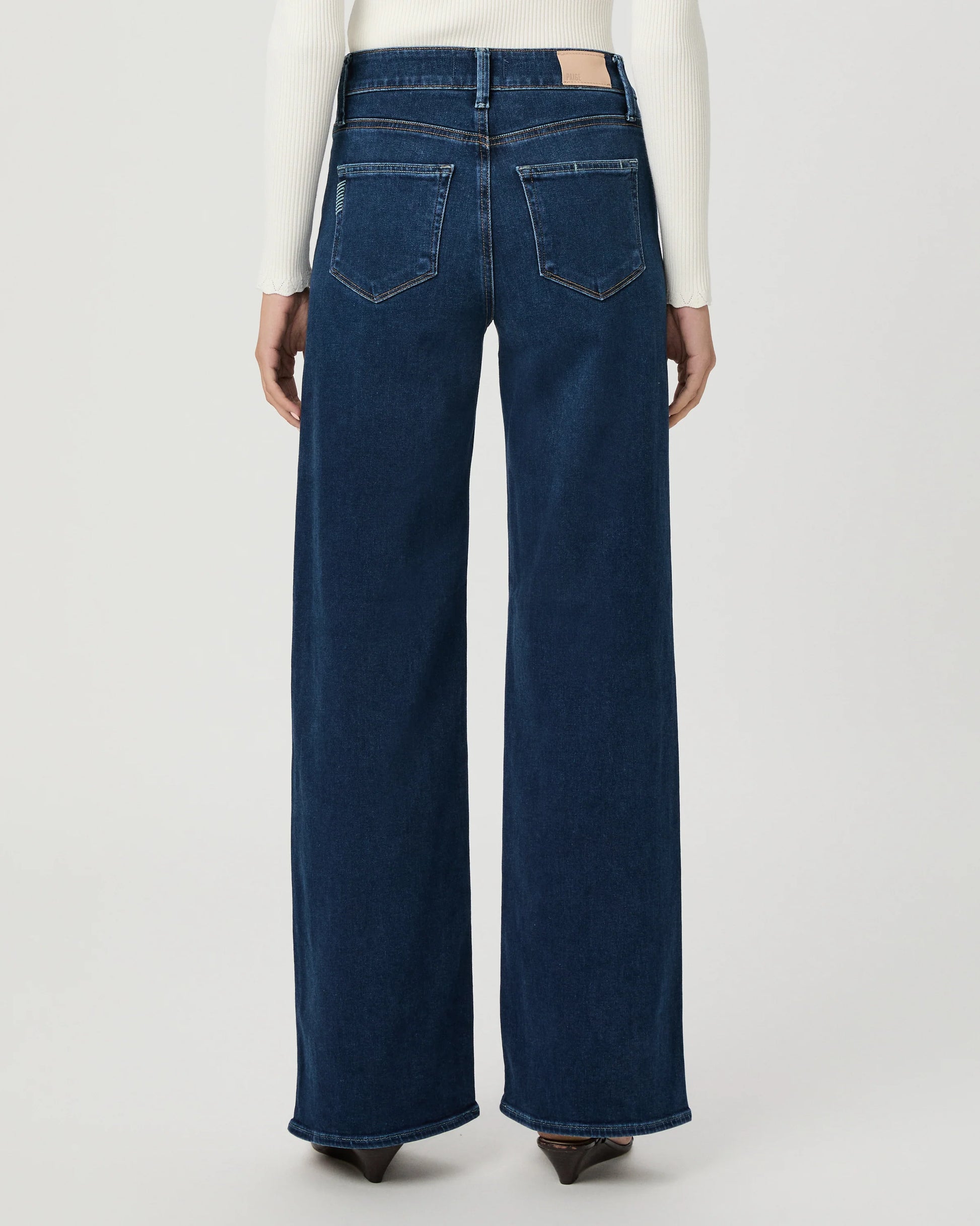 Back view of a model wearing dark blue wide-leg denim jeans with a white long-sleeve top, shown against a light gray studio background.