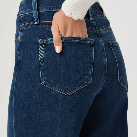 Close-up back view of a person wearing dark blue denim jeans with a hand in the back pocket, paired with a white ribbed long-sleeve top against a light gray background.
