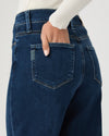 Close-up back view of a person wearing dark blue denim jeans with a hand in the back pocket, paired with a white ribbed long-sleeve top against a light gray background.