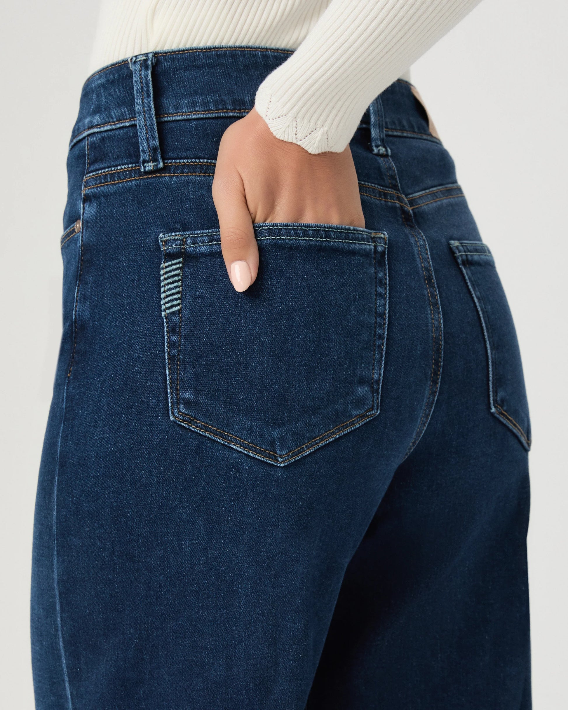 Close-up back view of a person wearing dark blue denim jeans with a hand in the back pocket, paired with a white ribbed long-sleeve top against a light gray background.