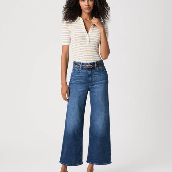 A woman modeling a cream and navy striped short-sleeve polo shirt paired with high-waisted, wide-leg blue jeans, a brown belt, and brown strappy sandals against a light gray studio background.