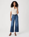 A woman modeling a cream and navy striped short-sleeve polo shirt paired with high-waisted, wide-leg blue jeans, a brown belt, and brown strappy sandals against a light gray studio background.
