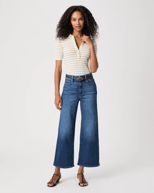 A woman modeling a cream and navy striped short-sleeve polo shirt paired with high-waisted, wide-leg blue jeans, a brown belt, and brown strappy sandals against a light gray studio background.