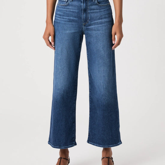 A model wearing a pair of high-waisted, wide-leg cropped blue denim jeans with a faded wash, paired with brown strappy sandals and a striped top, against a plain white studio background.