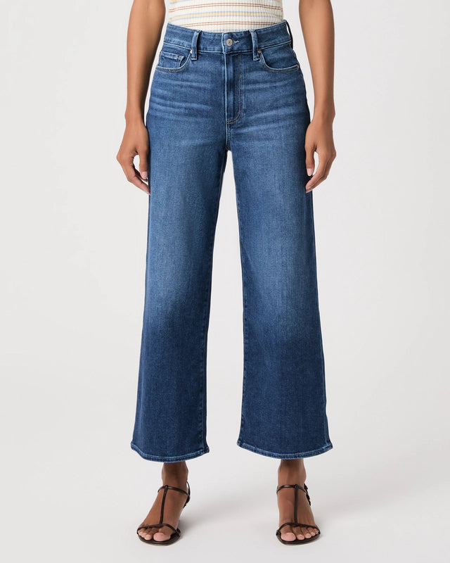 A model wearing a pair of high-waisted, wide-leg cropped blue denim jeans with a faded wash, paired with brown strappy sandals and a striped top, against a plain white studio background.