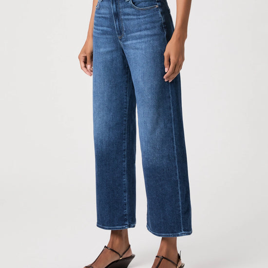 A model wearing a pair of high-waisted, wide-leg cropped blue denim jeans with brown wedge sandals, shown from the waist down on a light gray studio background.