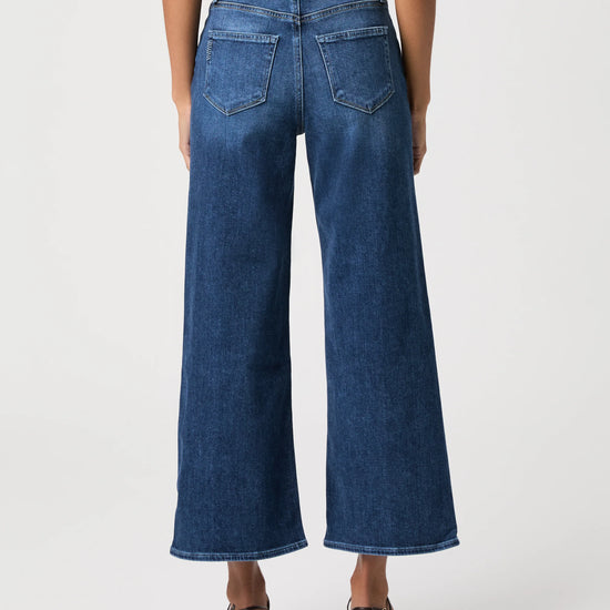 A model shown from the back wearing a pair of wide-leg cropped blue denim jeans, paired with a striped top and dark brown heeled sandals, against a light gray studio background.