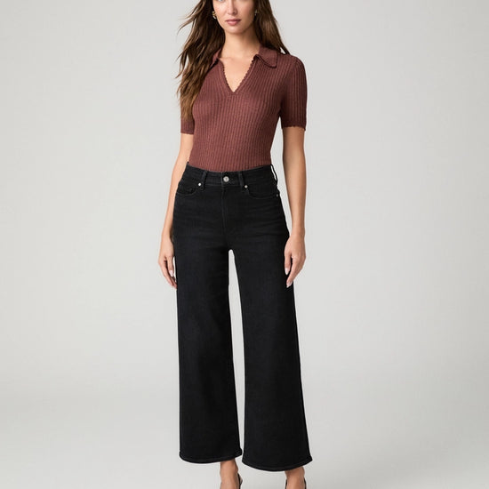 A woman modeling a short-sleeve ribbed knit polo shirt in a muted reddish-brown color paired with black wide-leg cropped jeans and black pointed heels, shot against a light gray studio background.