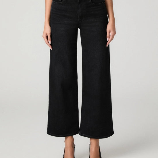 A model wearing a pair of black wide-leg cropped jeans with a ribbed top and black pointed-toe heels, photographed against a light gray studio background.
