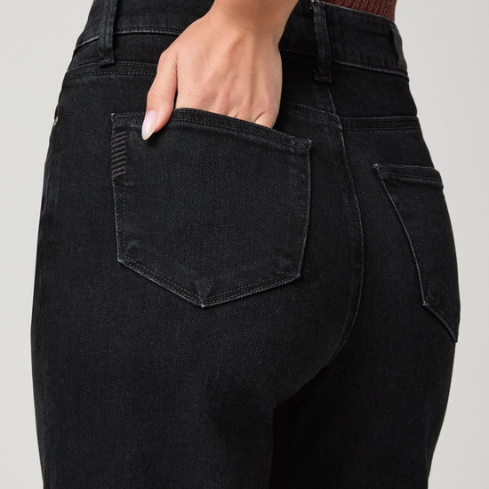 Close-up back view of a person wearing black high-waisted jeans, with one hand tucked into the back pocket, against a light gray background.