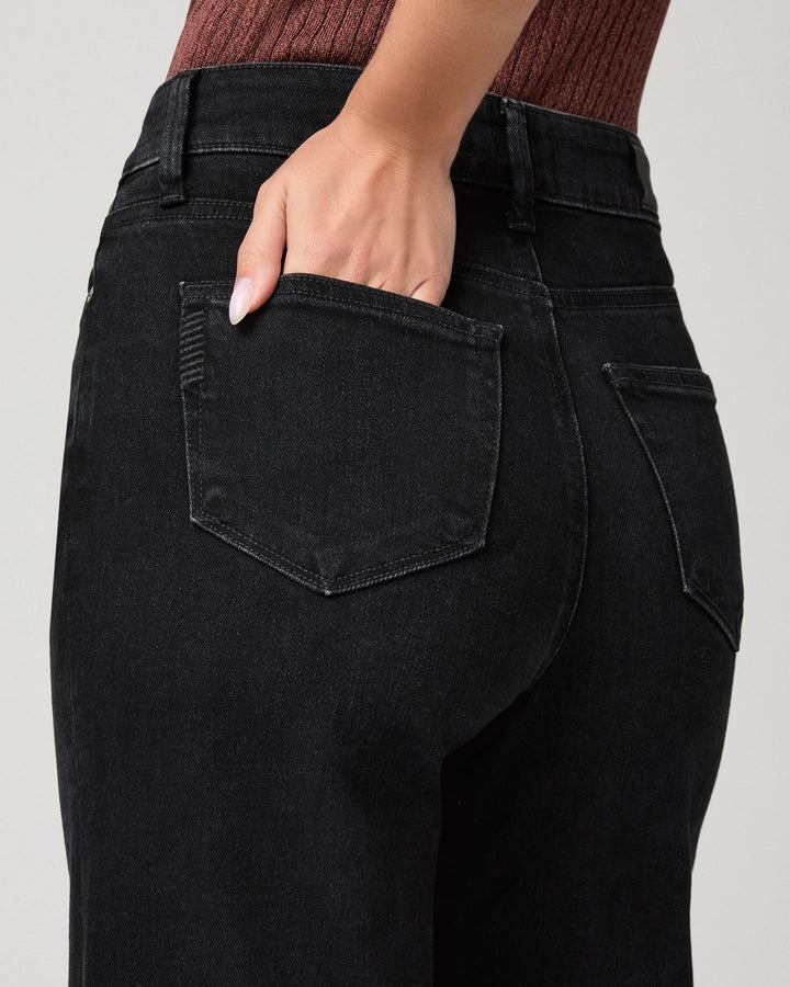 Close-up back view of a person wearing black high-waisted jeans, with one hand tucked into the back pocket, against a light gray background.