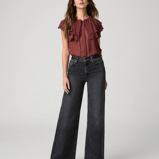 A woman modeling a dark brown ruffled sleeveless blouse paired with wide-leg dark gray jeans and black pointed-toe heels, photographed against a light gray studio background.