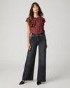 A woman modeling a dark brown ruffled sleeveless blouse paired with wide-leg dark gray jeans and black pointed-toe heels, photographed against a light gray studio background.