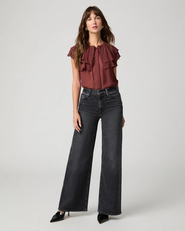 A woman modeling a dark brown ruffled sleeveless blouse paired with wide-leg dark gray jeans and black pointed-toe heels, photographed against a light gray studio background.