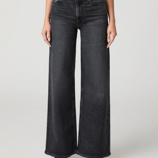 A model wearing a pair of wide-leg dark gray denim jeans, paired with a rust-colored top and black pointed shoes, photographed against a neutral light gray studio background.