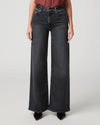 A model wearing a pair of wide-leg dark gray denim jeans, paired with a rust-colored top and black pointed shoes, photographed against a neutral light gray studio background.