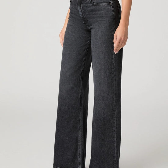 A model wearing a pair of dark gray wide-leg denim jeans, paired with a burgundy top and black pointed-toe heels, shown from the waist down on a light gray studio background.
